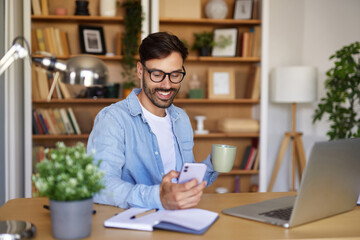 Fototapeta premium A man sits at a desk in a home office, smiling as he holds a smartphone in one hand and a cup in the other. Books and plants decorate the warm space with natural light streaming in.