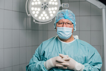Experienced male surgeon in blue surgical attire, wearing glasses and a mask, stands confidently in...