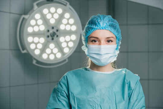 Female surgeon in blue surgical attire and mask, standing confidently in a well-lit operating room, ready for a procedure, showcasing professionalism and dedication to patient care