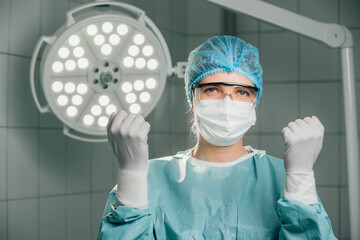 Female surgeon in green scrubs and surgical mask, confidently raising hands in operating room,...