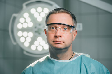 Male surgeon in surgical scrubs and protective eyewear, standing confidently in a bright operating...