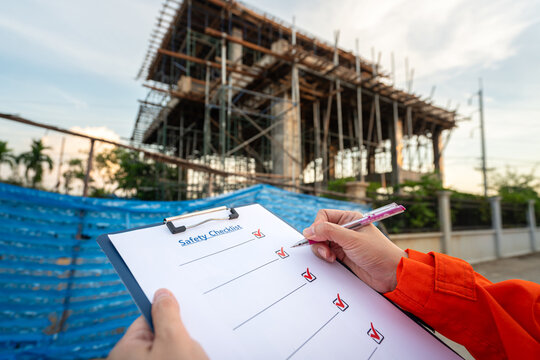 Action of an engineer is checkling on safety checklist form at construction working site (with background of house builing). Industrial safe work practice concept, close-up with selective focus.