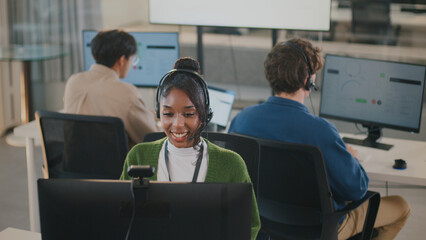 Cheerful customer service representative wearing headset is working at computer in modern office environment, with two colleagues in background also engaged in similar tasks