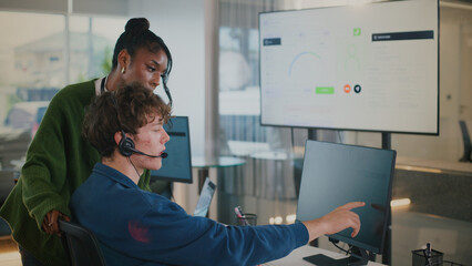 Focused customer service representative wearing headset is working computer while colleague leans to assist, both collaborating modern office environment with screens displaying data background