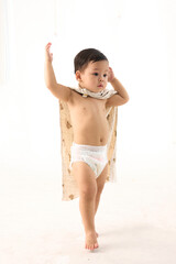 Toddler boy walking forward with blanket wrapped around shoulders, white background