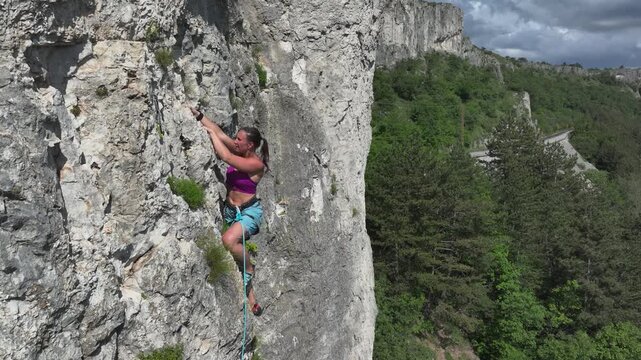 AERIAL: Female athlete climbing a rock face with a backdrop of a lush green forest and a scenic mountain railroad. She is focused on her next move while ascending a rocky outcrop in a scenic location.