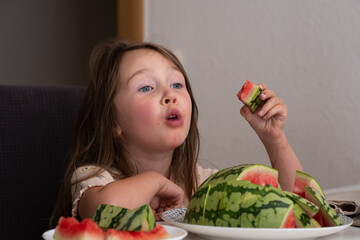 A small beautiful Caucasian girl sits at the table and eats a watermelon, making funny faces. There...