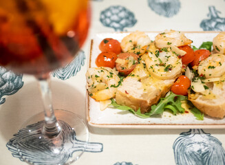 Plate of shrimp and tomatoes sits on a table next to a wine glass. The wine glass is half full and the plate of food is almost empty. Scene is light and casual
