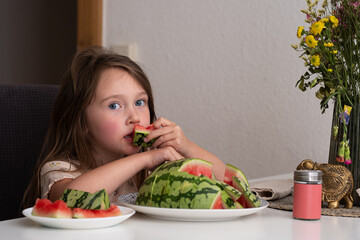 A small beautiful Caucasian girl sits at the table and eats a watermelon, making funny faces. There...