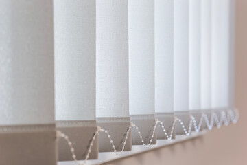 Close-up of vertical blinds with textured fabric panels in light beige. Bottom weights connected by...