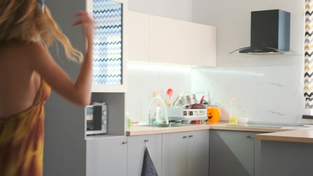 Blonde girl twirling and dancing in headphones at home, immersed in music. She moves freely in a flowing dress, enjoying the rhythm in a bright modern kitchen. Youthful and full of energy and freedom.