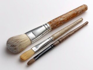 Fototapeta premium Three paintbrushes of varying sizes and bristle types, with wooden handles showing age and use, rest on a plain white background