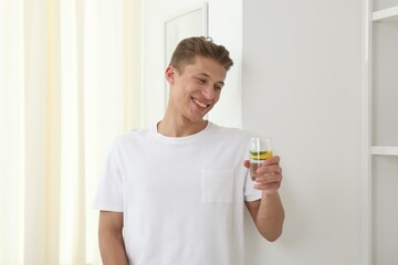Smiling man with glass of water with cucumber and lemon near white wall indoors. Refreshing drink
