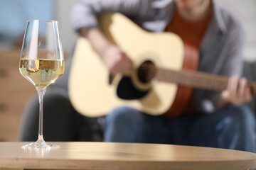 Glass with white wine on table and man playing guitar indoors, selective focus