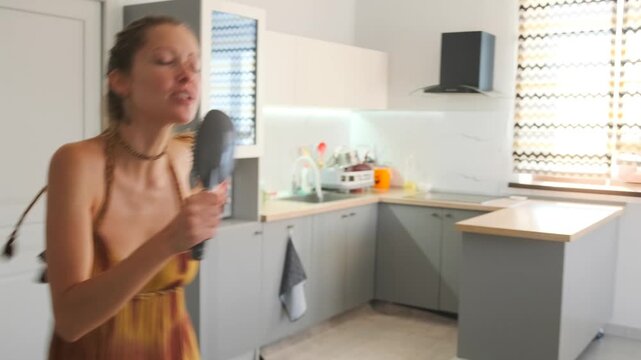 Young woman in a boho summer dress sings into a hairbrush and dances playfully in a bright modern kitchen. Joyful, candid, and carefree morning moment at home.