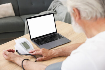 Senior man measuring blood pressure while having online consultation with doctor via laptop at table indoors, closeup