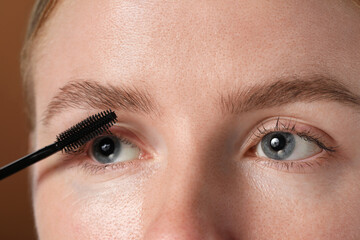 Young woman applying mascara onto her eyelashes on brown background, closeup