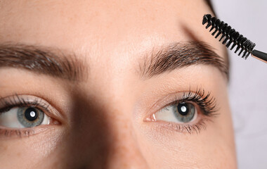 Young woman brushing her eyebrow with spoolie on light grey background, closeup