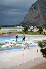 Community members spray-paint vibrant mural on basketball court by the beach as storm clouds gather...