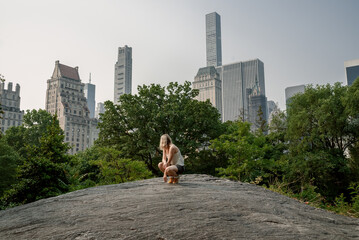 Woman climbing on a large rock in Central Park with skyscrapers in the background on a sunny day