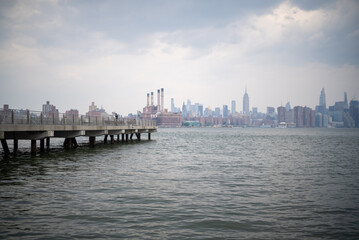 Naklejka premium Pier extends into the water while city skyline looms under cloudy sky in New York during late afternoon