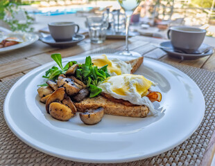 Gourmet breakfast of Eggs Benedict with sauteed mushrooms and greens, served al fresco on a hotel terrace by the swimming pool.Luxury vacation brunch.Perfect summer holiday morning