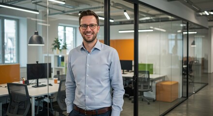 Fototapeta premium A man smiles in front of glasswalled offices wearing glasses and a buttondown shirt
