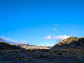 A vast, panoramic landscape of a wide valley under a bright blue sky with a few scattered white clouds. The valley is enclosed by a series of rolling hills and mountains on either side