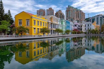 Reflective cityscape featuring colorful buildings bordering water with distant highrises under a blue sky