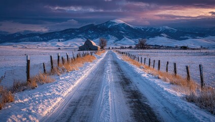 Snow-covered rural road leads to a rustic barn nestled in a valley, mountains and dramatic sunset in the background