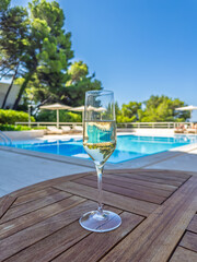 Glass of champagne on a wooden table by a swimming pool at a luxury resort.Summer vacation and relaxation concept. Flute of chilled prosecco or champagne with blurred swimming pool in the background
