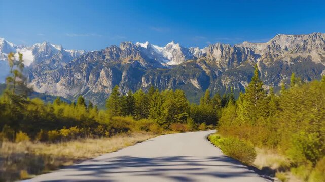 Beautiful winding road through lush green forest, leading towards majestic snow-capped mountains under a bright blue summer sky.