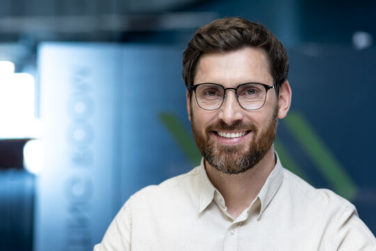 Close-up portrait of a young man in glasses and a shirt who is in the office, smiling and looking at the camera
