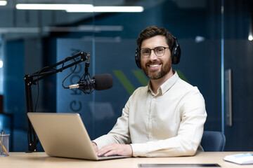 Portrait of a young smiling man sitting at a desk in the office, wearing headphones in front of a...