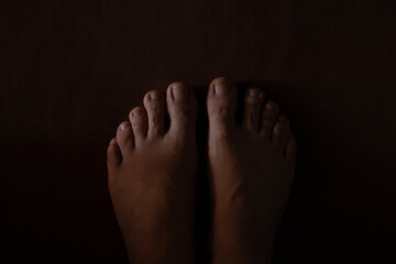 Fototapeta premium Female bare feet standing on brown floor in semi-darkness in apartment