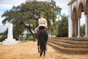 Young female horsewoman riding a black Spanish thoroughbred horse, trotting with him, seen from the back. She has a saddle on the side and a riding crop. Concept horse riding, horsewoman, dressage.