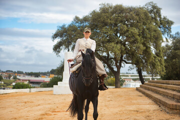 Young female horsewoman riding a black Spanish thoroughbred horse, trotting with him towards the...