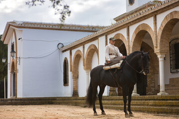 Young horsewoman riding a black Spanish thoroughbred horse next to an old hermitage. She has a...