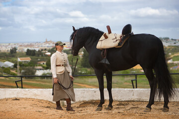 Young woman rider, posing next to her black Spanish thoroughbred horse, which she is holding by the...