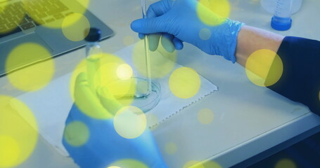 Stirring gloved hand transferring liquid in glass container on laboratory bench, with laptop