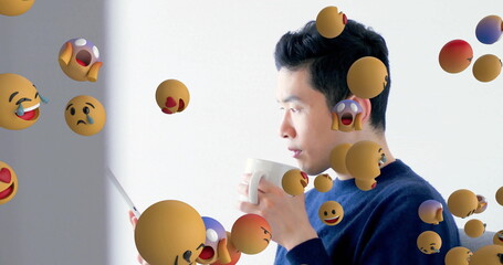 Standing Asian man holding tablet and mug in white room, with floating emoji spheres, copy space