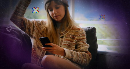 Sitting woman wearing knit sweater, beanie scrolling smartphone on dark sofa by large paned window