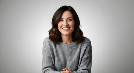 Happy young woman with shoulder-length dark hair and a bright smile wearing a cozy gray sweater sitting against a plain light gray background for lifestyle and fashion stock photography