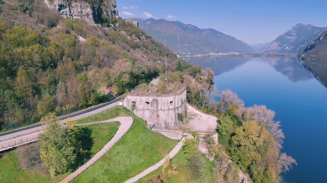 Aerial shot of Lake Idro in front of Rocca d'Anfo which is nestled on forested mountain with a road