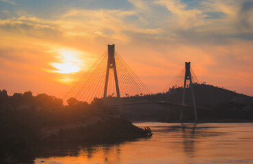 Obraz premium The silhouette of a majestic suspension bridge over a calm river at sunset, with a vibrant orange and blue sky. The golden hour creates a peaceful and dramatic atmosphere.