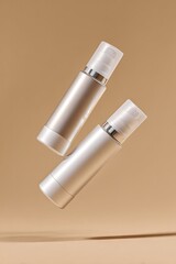 Two sleek, cylindrical cosmetic bottles, one champagne gold, one silver, levitate against a muted beige backdrop.  Minimalist product photography with subtle shadows