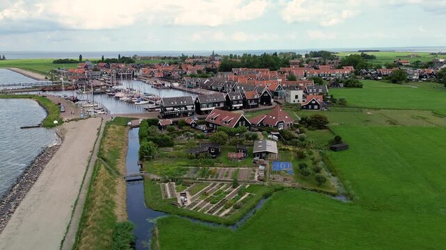 Aerial view of a charming Dutch harbor town with traditional red-roofed houses, moored sailboats, and calm waters surrounded by green fields and open sky, creating a serene coastal scene.