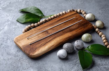 Two slender, silver-toned metal rods rest on a dark brown wooden board, surrounded by smooth stones, wooden beads, and vibrant green leaves on a textured gray surface
