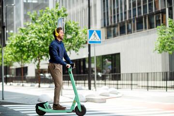 Young businessman on electric scooter listening to music through headphones riding in city during...