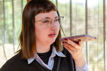 Portrait of young female employee or student wearing glasses sending audio message with a mobile...
